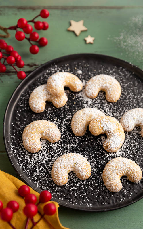 Klassische Vanillekipferl mit Puderzucker auf einem schwarzen Teller, weihnachtlicher Hintergrund 